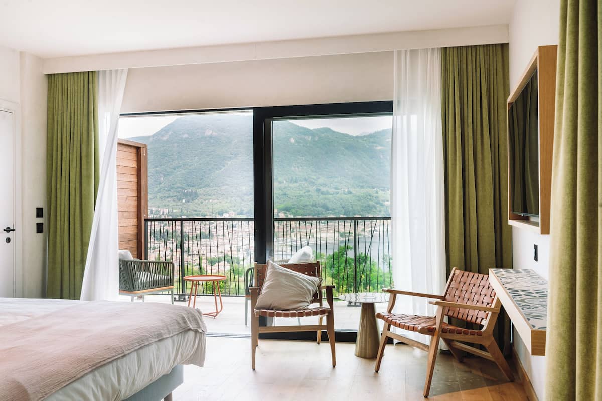 A hotel room with a bed, a chair, a small table, and a balcony with a view of mountains.