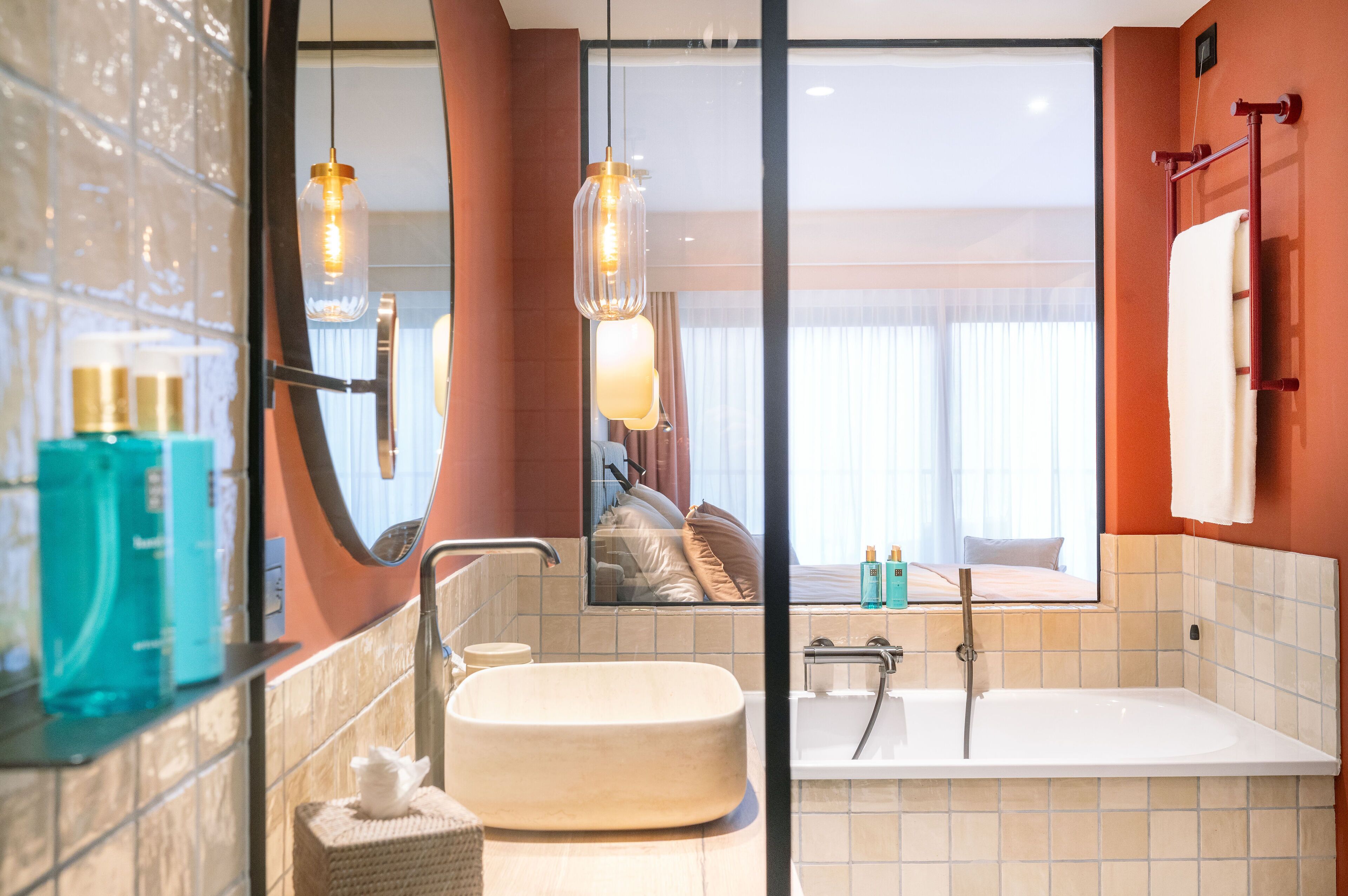 A modern bathroom with a large sink, a bathtub, and a mirror reflecting a bedroom with a bed and curtains.