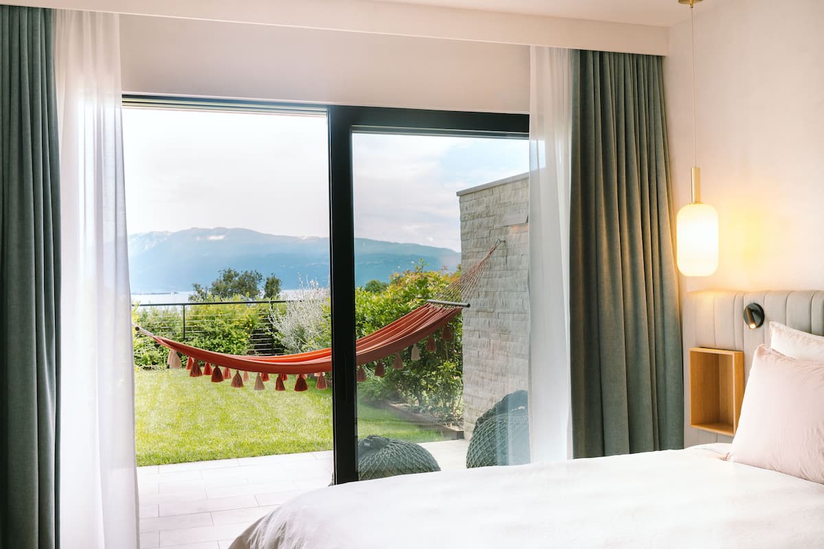 A hotel room with a large window, a bed, a nightstand, and a view of a garden and mountains.