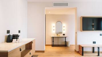 A hotel room with a wooden floor, a white vanity with a mirror and lights, a coffee maker, and a TV mounted on the wall.