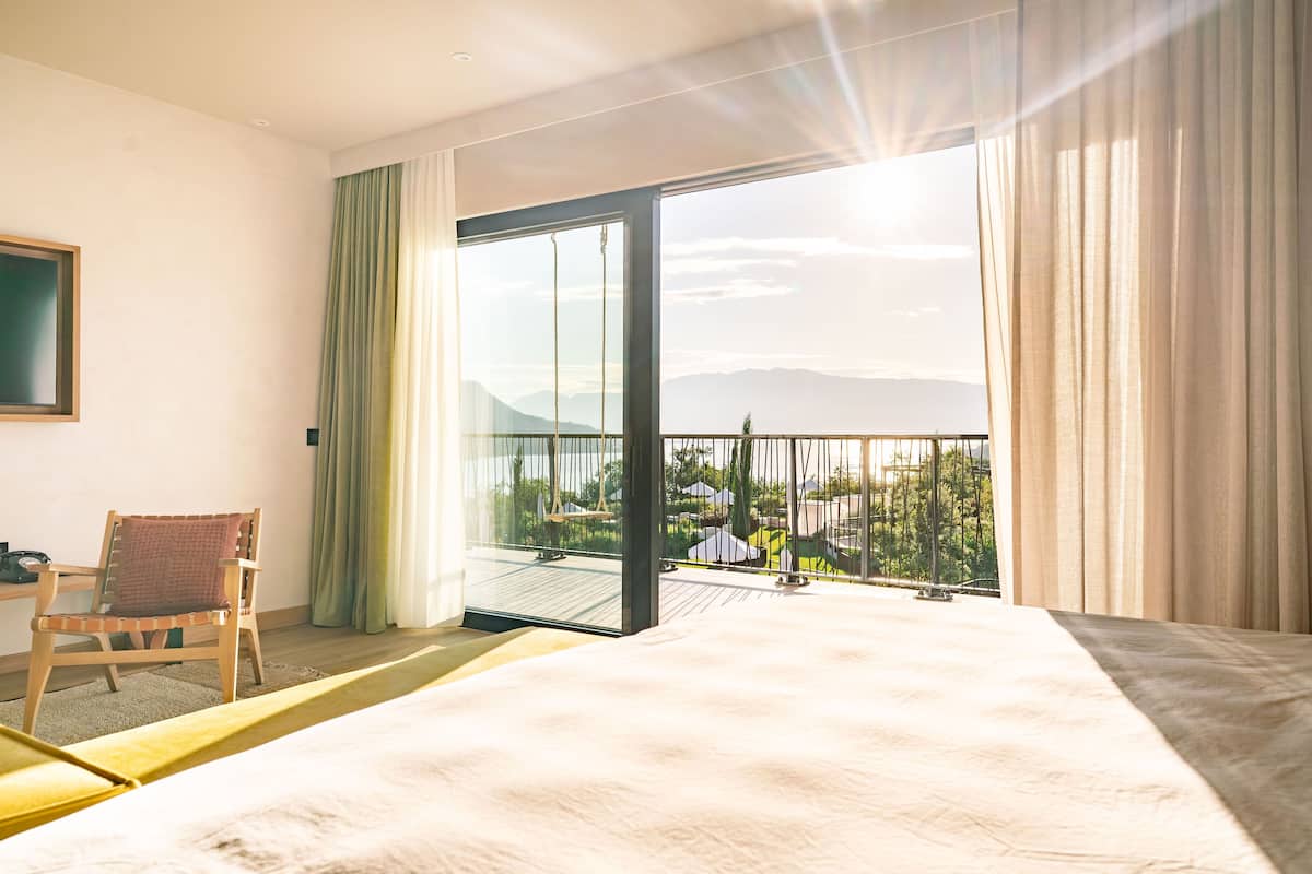 A hotel room with a large bed, a chair, a desk, and a balcony with a view of mountains and greenery.