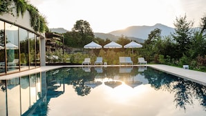 A modern pool area with clear glass walls, lounge chairs, and umbrellas, surrounded by lush greenery and mountains. - A-ROSA Gardasee (Salò)