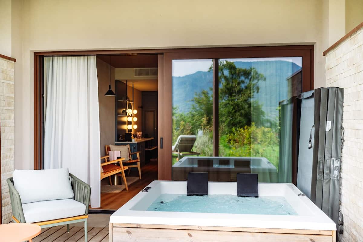 A room with a hot tub, a chair, a table, and a view of mountains.