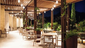 An outdoor patio with wooden tables and chairs, surrounded by potted plants and a brick wall.