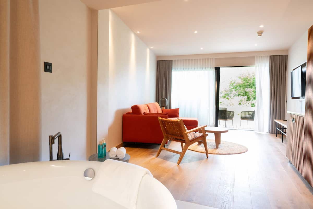 A modern hotel room with a large bed, a red sofa, a wooden chair, a small table, and a TV mounted on the wall.
