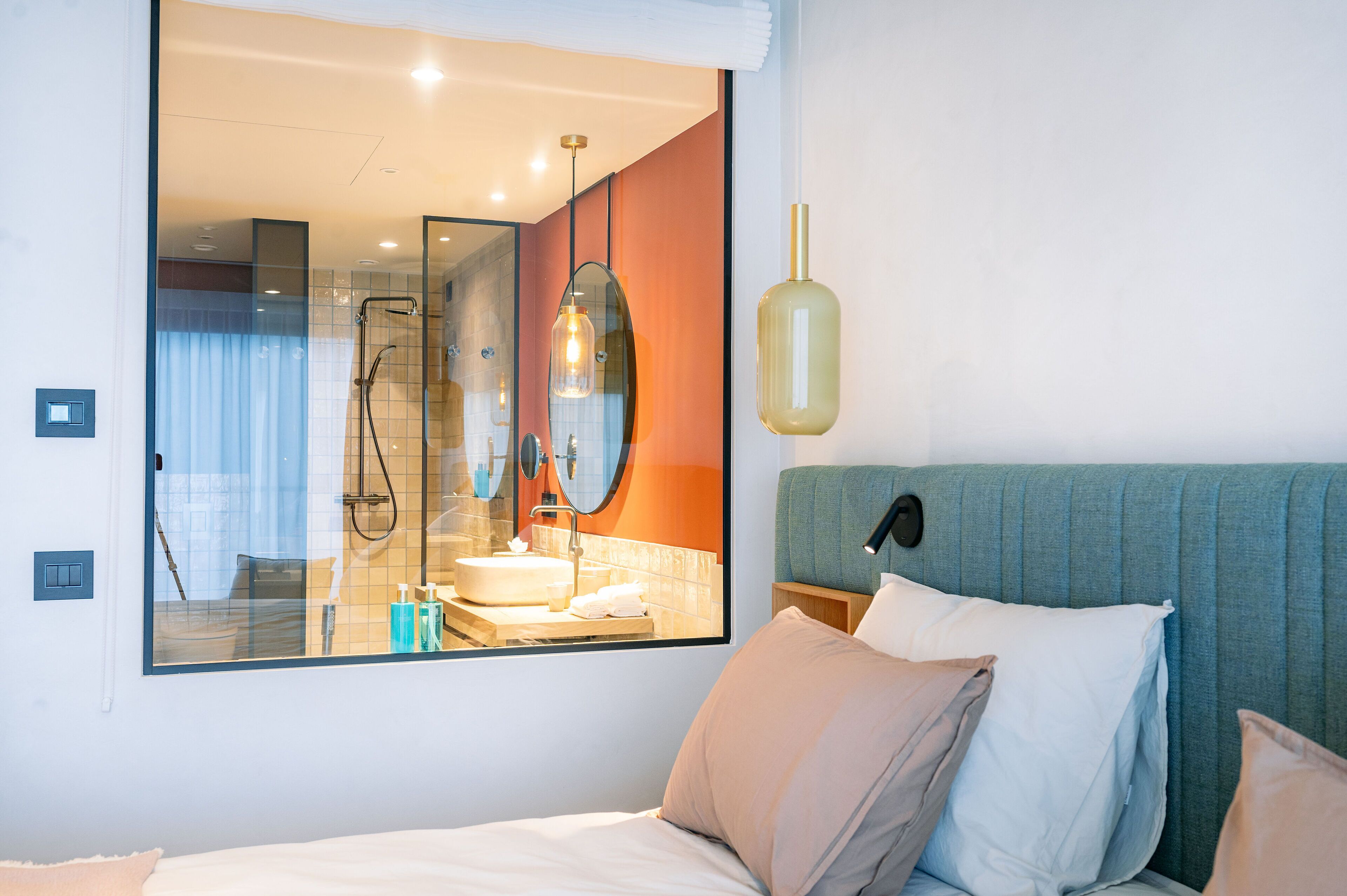 A modern hotel room with a large mirror reflecting the bathroom, a bed with multiple pillows, and a unique pendant light.