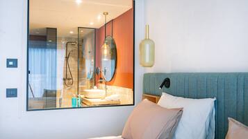 A modern hotel room with a large mirror reflecting the bathroom, a bed with multiple pillows, and a unique pendant light.