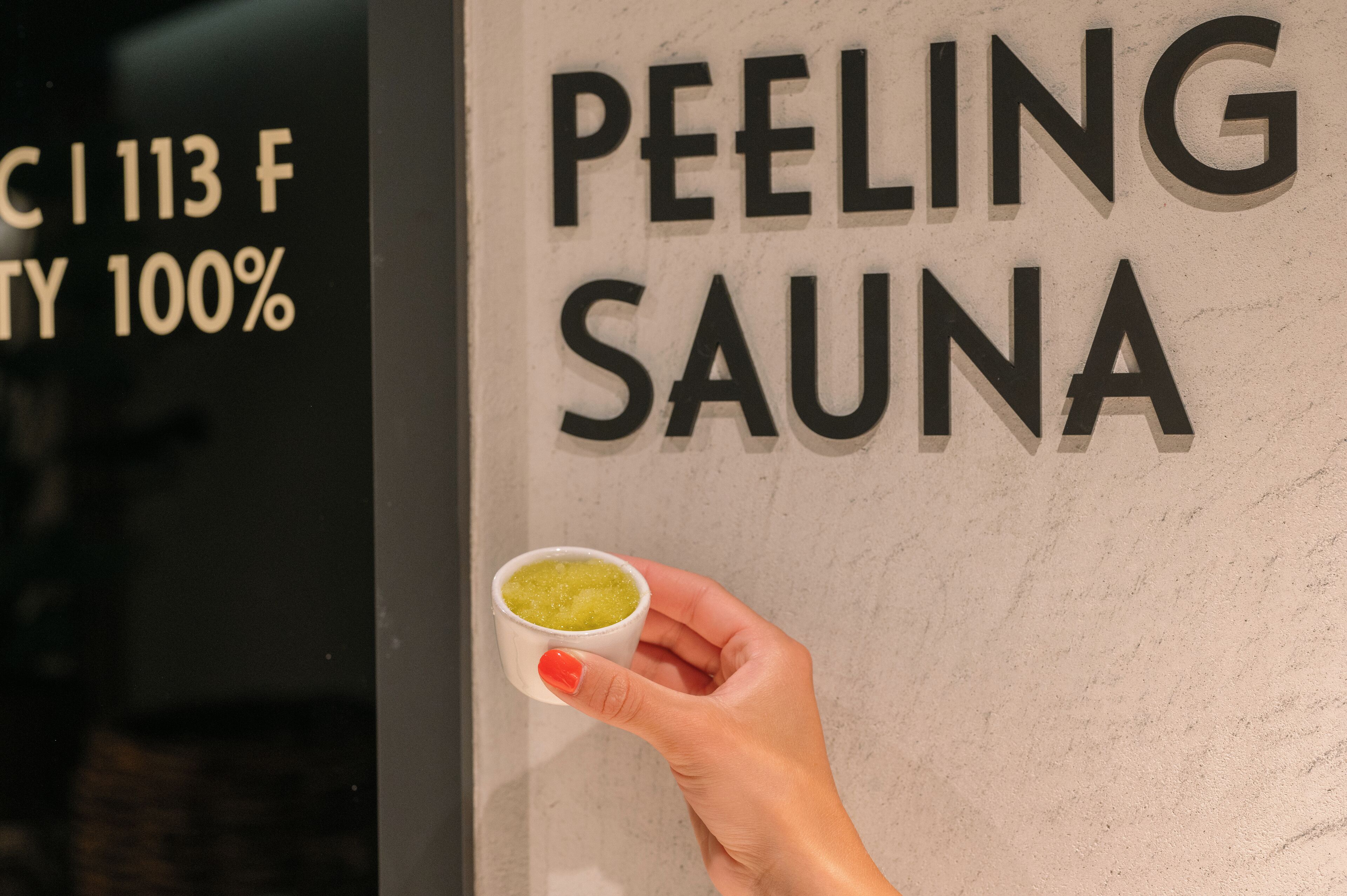 Una mano sostiene una taza pequeña con un líquido verde, frente a una pared donde se lee 'PEELING SAUNA'.