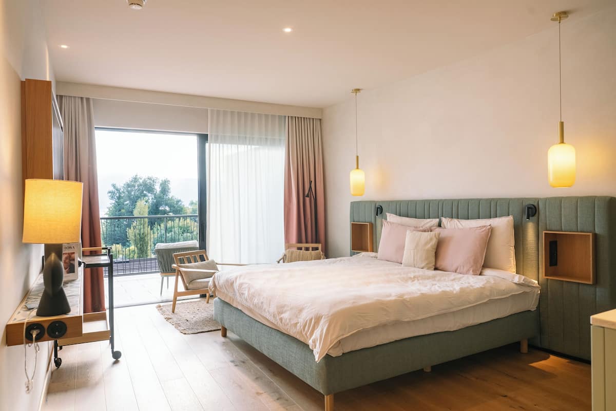 A bedroom with a bed, bedside tables, a desk, a chair, and a view through a large window.