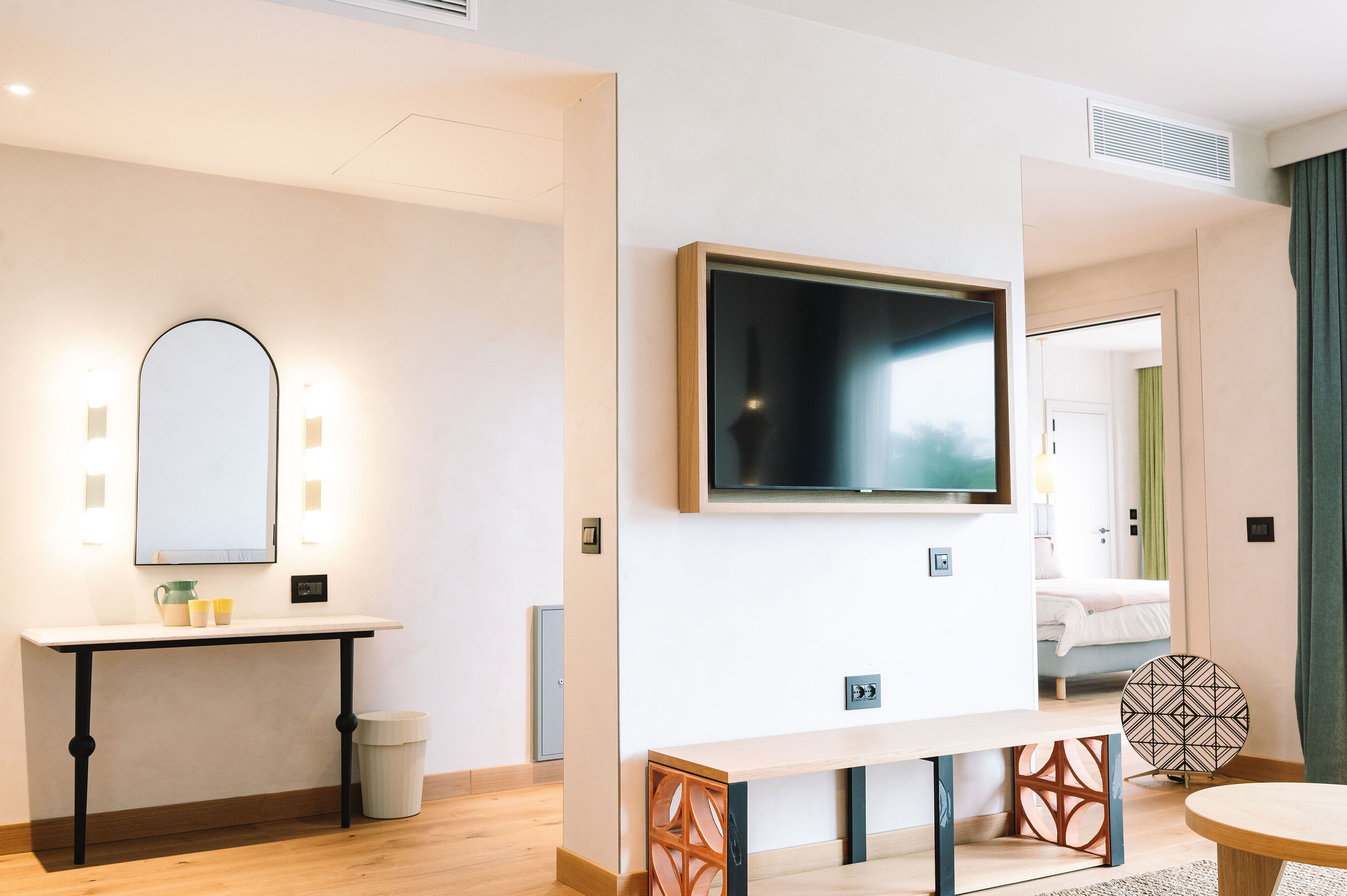 Una moderna camera d'hotel con una grande televisione a schermo piatto montata sulla parete, un comodino, una panca e uno specchio illuminato da applique.