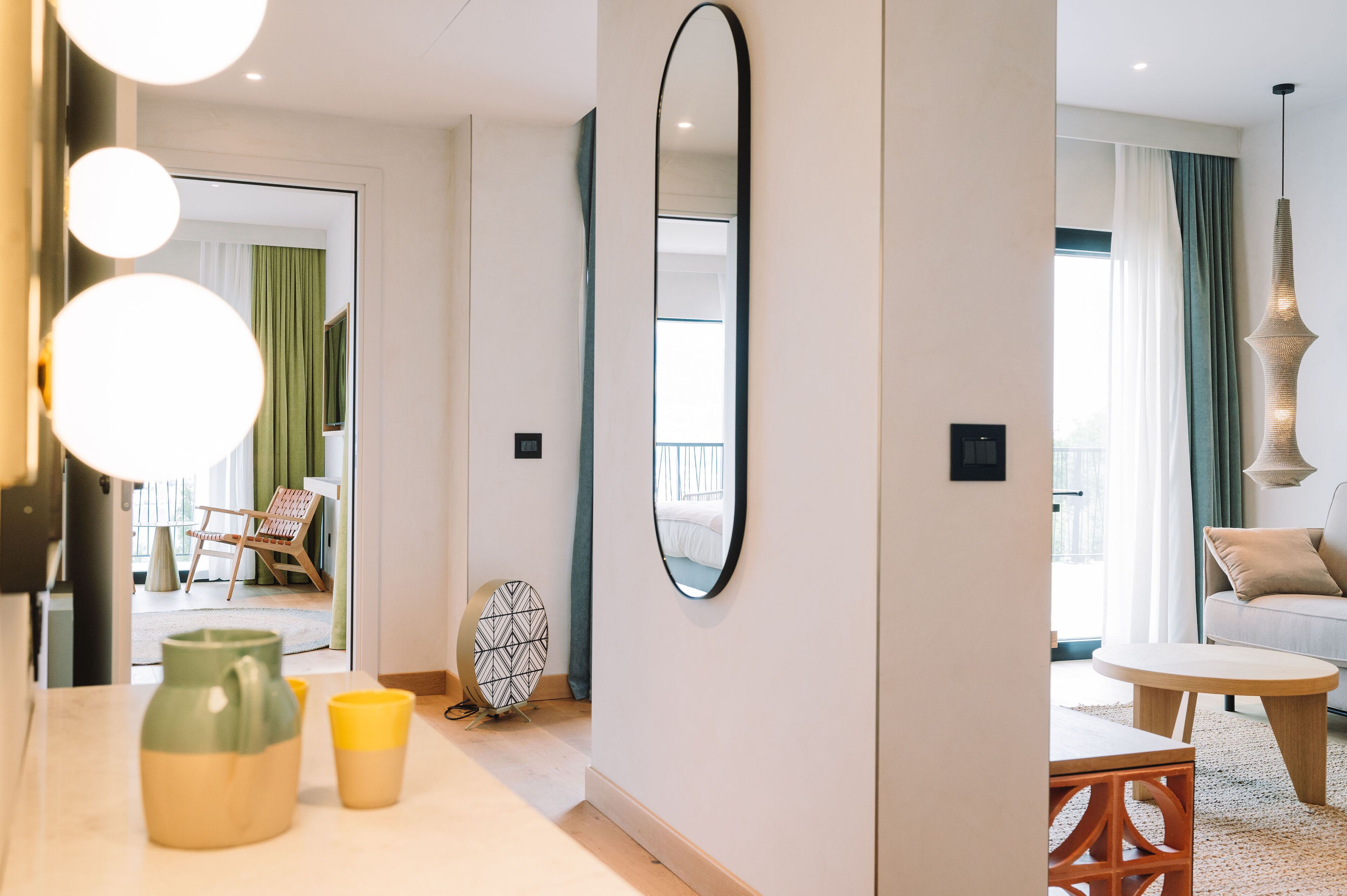 Une chambre d’hôtel moderne avec un grand miroir, une table ronde et un canapé. On peut admirer, depuis la chambre, un balcon avec une chaise et une table à l’extérieur.