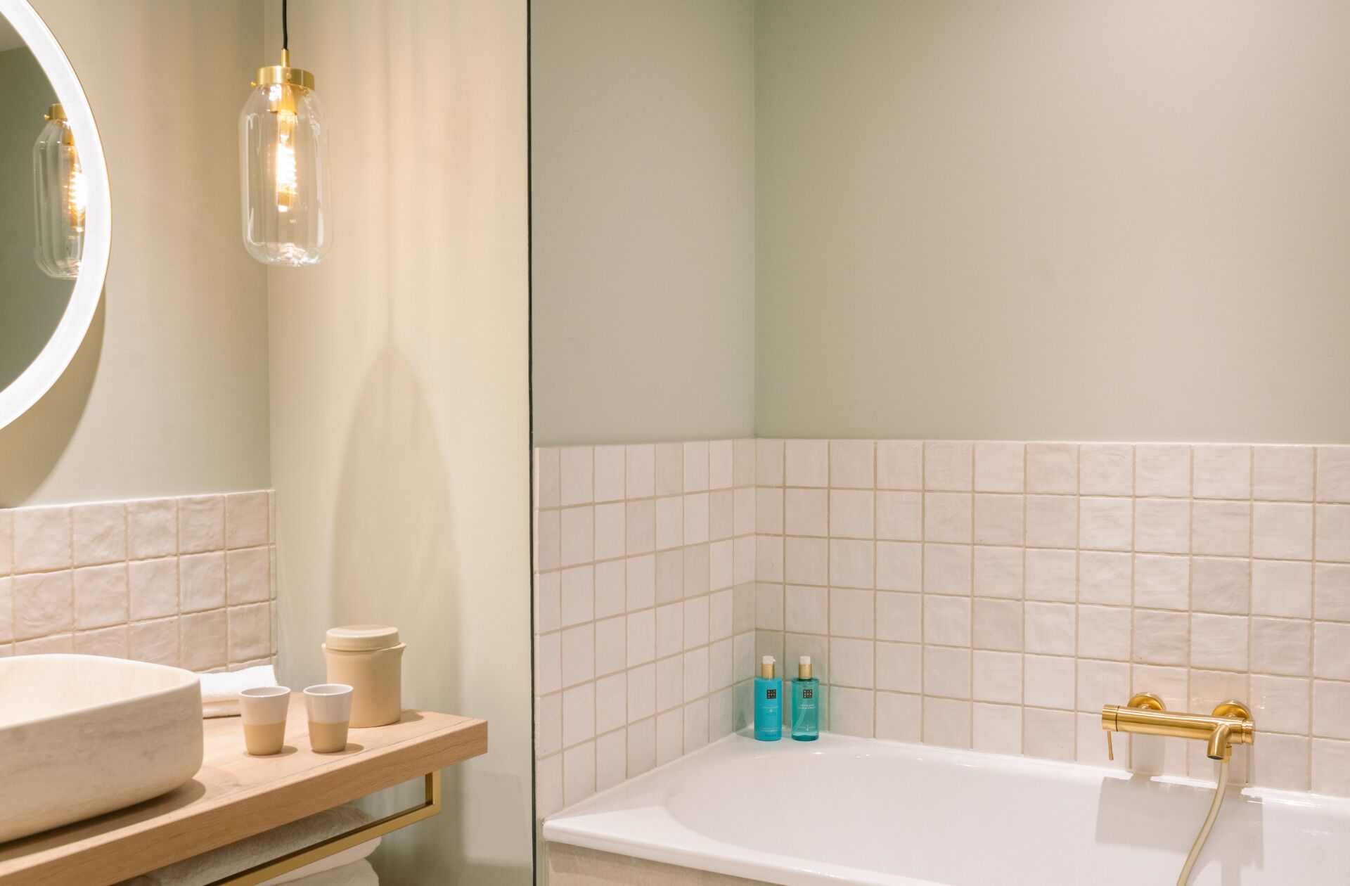 A bathroom with a round mirror, a wooden vanity, a white sink, a beige countertop, a white bathtub, and a shower with a gold faucet.