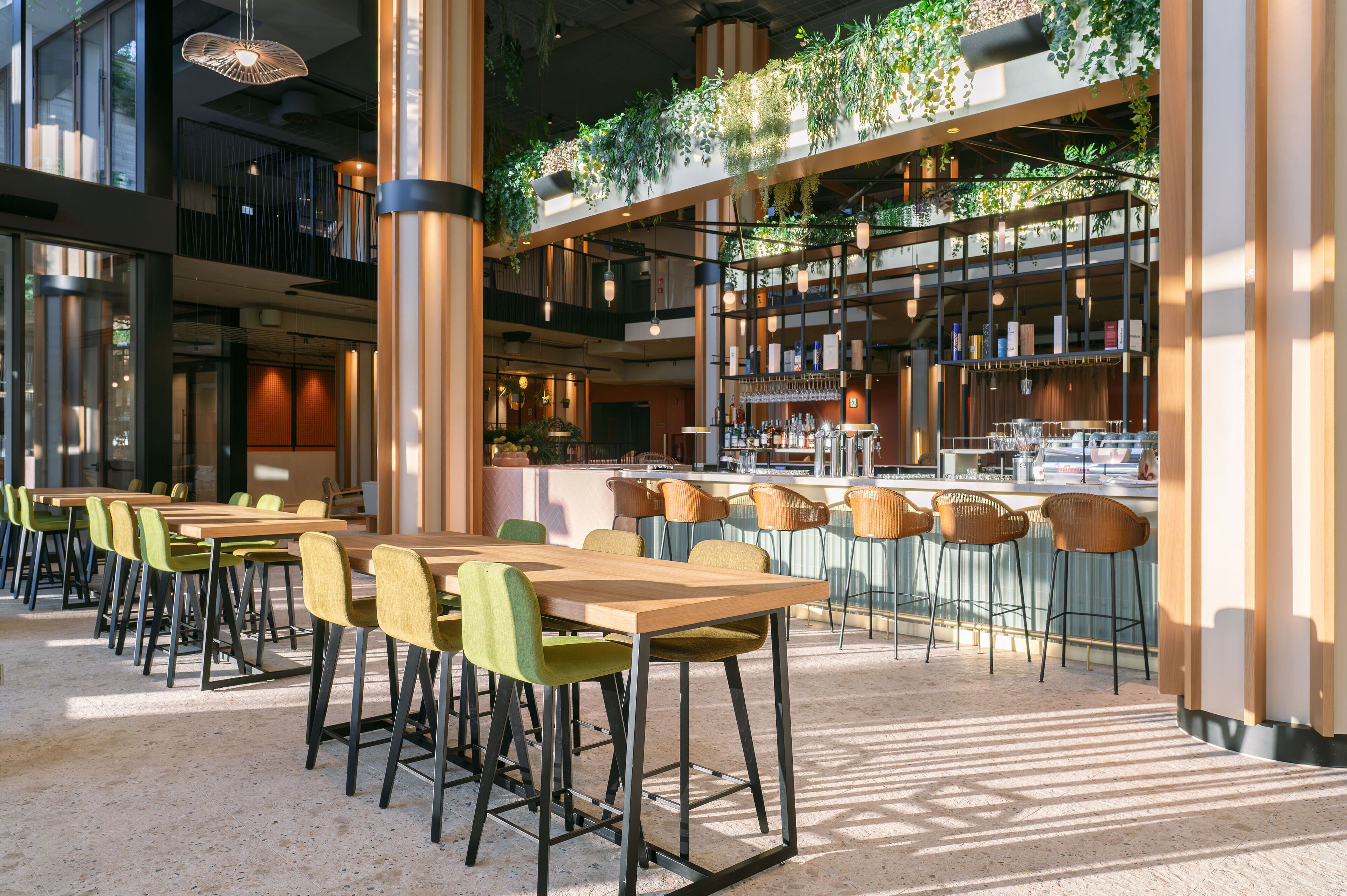 Un'area ristorazione moderna con diversi tavoli e sedie, un bancone bar con scaffali pieni di bottiglie e un soffitto decorato con piante pensili.