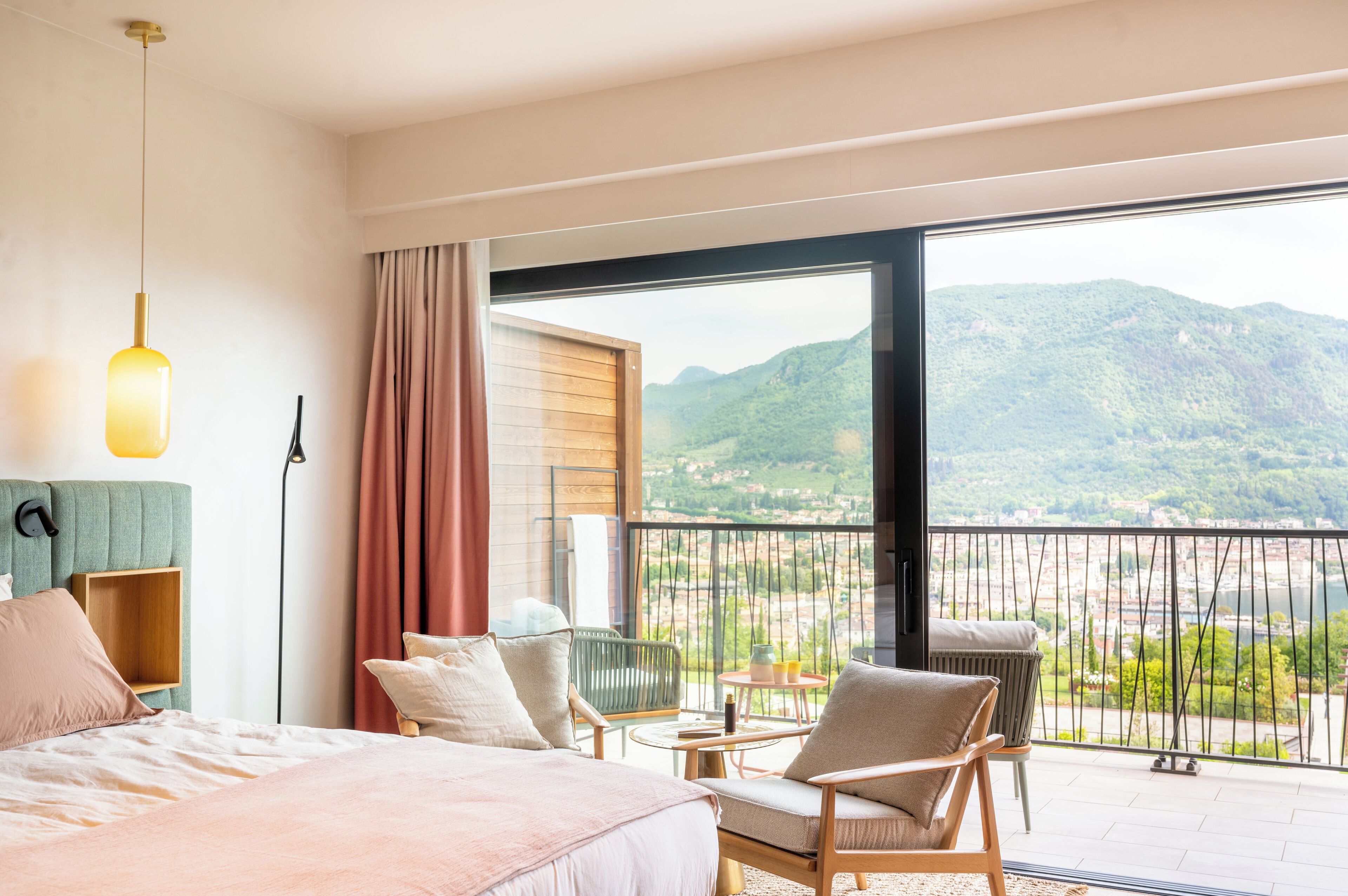 A hotel room with a bed, a chair, a small table, a lamp, and a balcony with a view of mountains and a river.