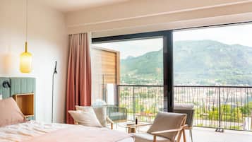 A hotel room with a bed, a chair, a small table, a lamp, and a balcony with a view of mountains and a river.
