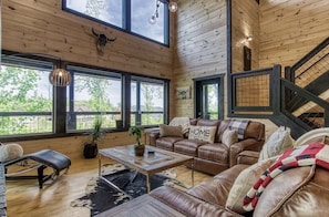 A spacious living room with a leather sofa, a coffee table, and a view of the outdoors through large windows. - ✨ Sunset:Beautiful Modern cabin in the Best Location 🏅 (Gatlinburg)