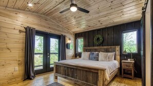 Ein Schlafzimmer in einer Holzhütte mit einem großen Bett, einem Deckenventilator und Blick ins Freie durch eine Schiebetür.