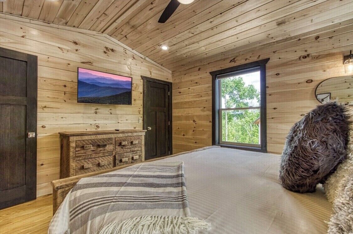 A wooden cabin bedroom with a bed, a dresser, a TV, and a window.