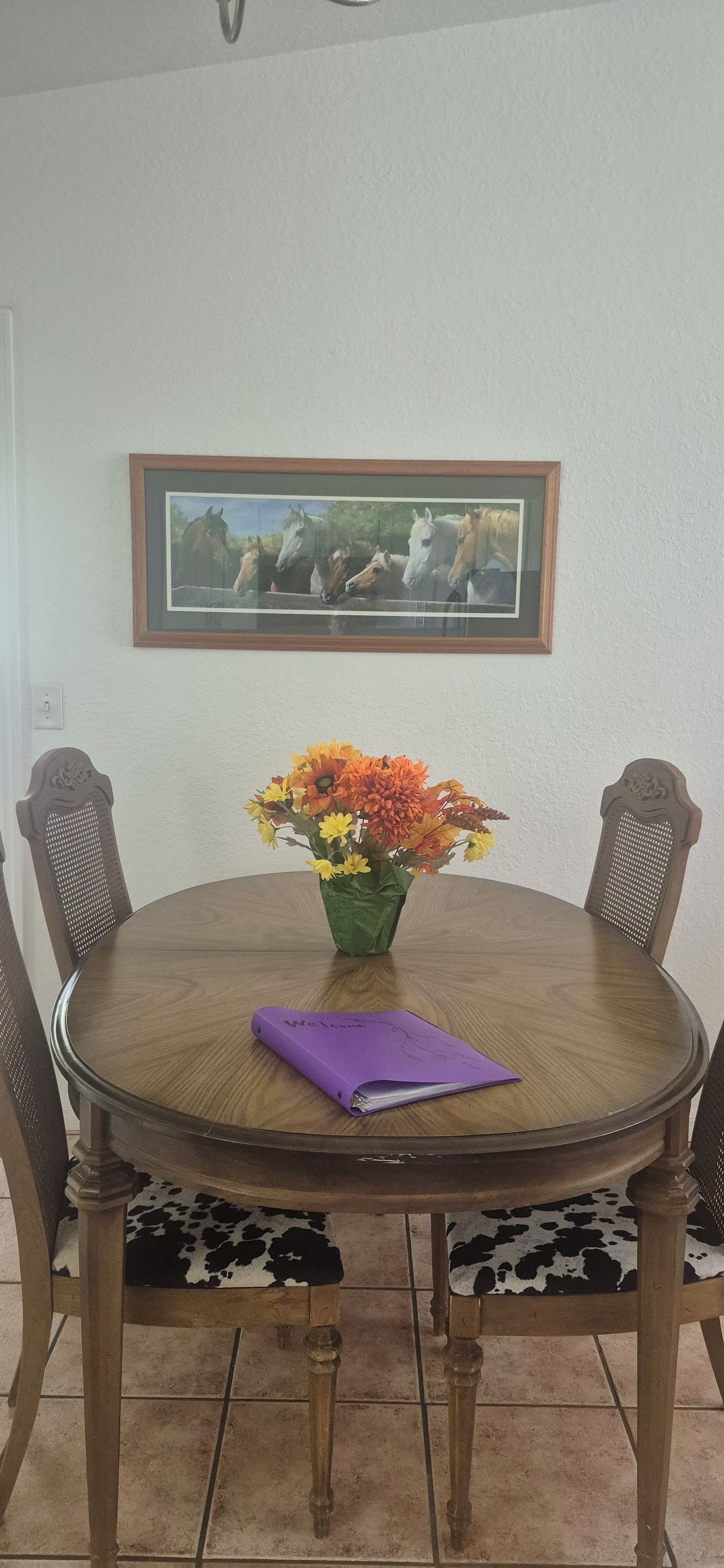 A round wooden dining table with a purple folder on it, surrounded by two chairs with a cow print cushion.