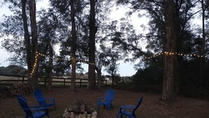 An outdoor seating area with three blue chairs arranged around a fire pit, surrounded by trees and a wooden fence.