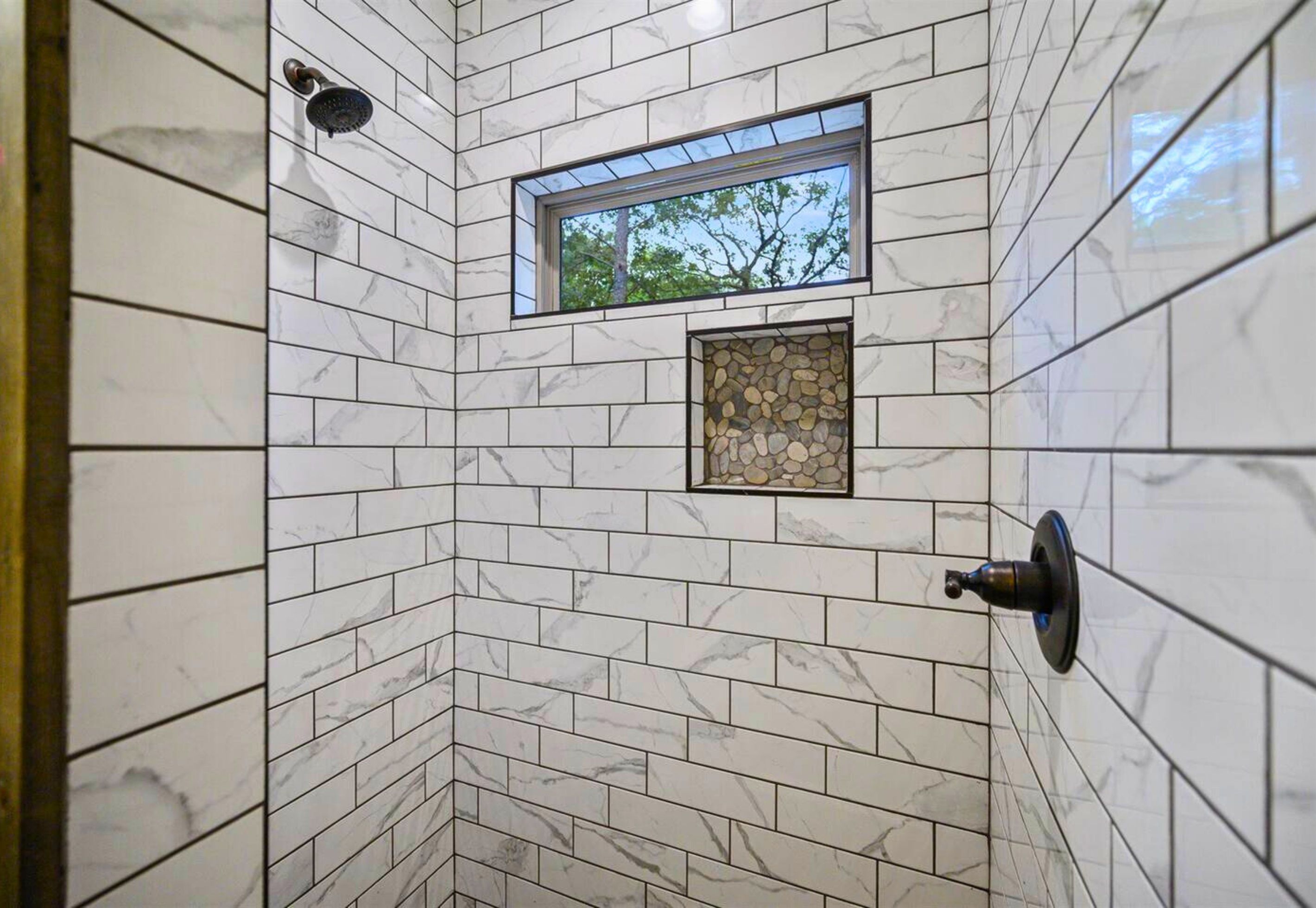 Une douche aux parois recouvertes de carreaux de marbre blanc, une fenêtre et un mur d’accent en pierre.