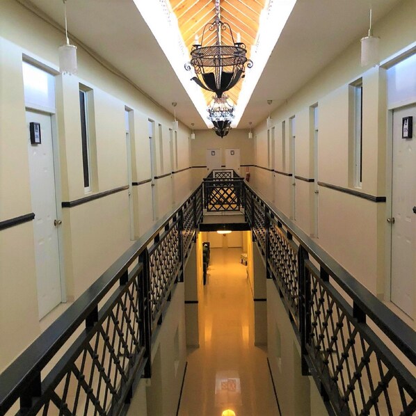 A hotel corridor with a central staircase, white walls, and a decorative chandelier.