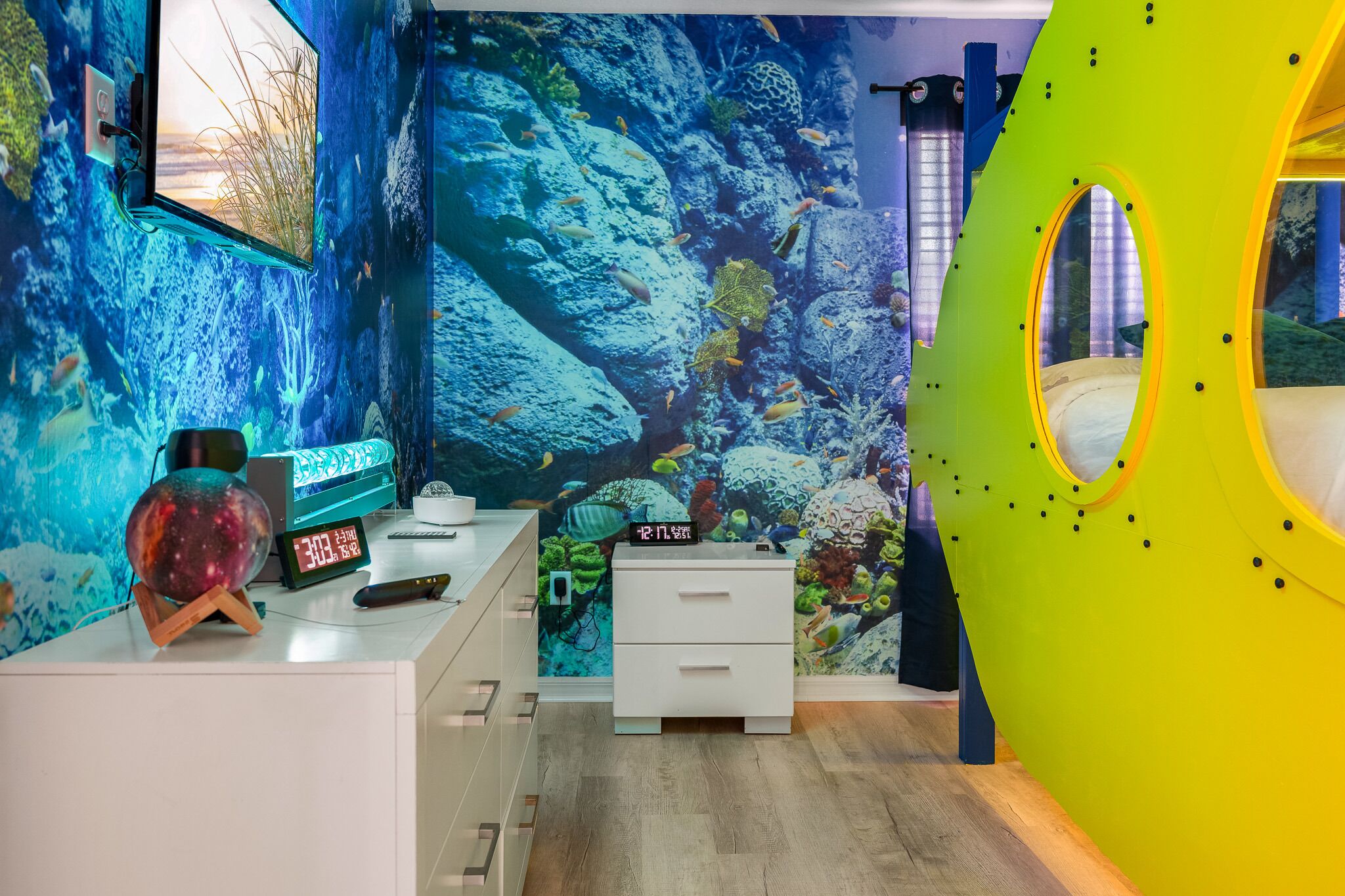 Ein Zimmer mit einer großen Aquariumwand, einem Bett mit rundem Fenster und einem weißen Nachttisch mit einer Uhr und Fernbedienung.