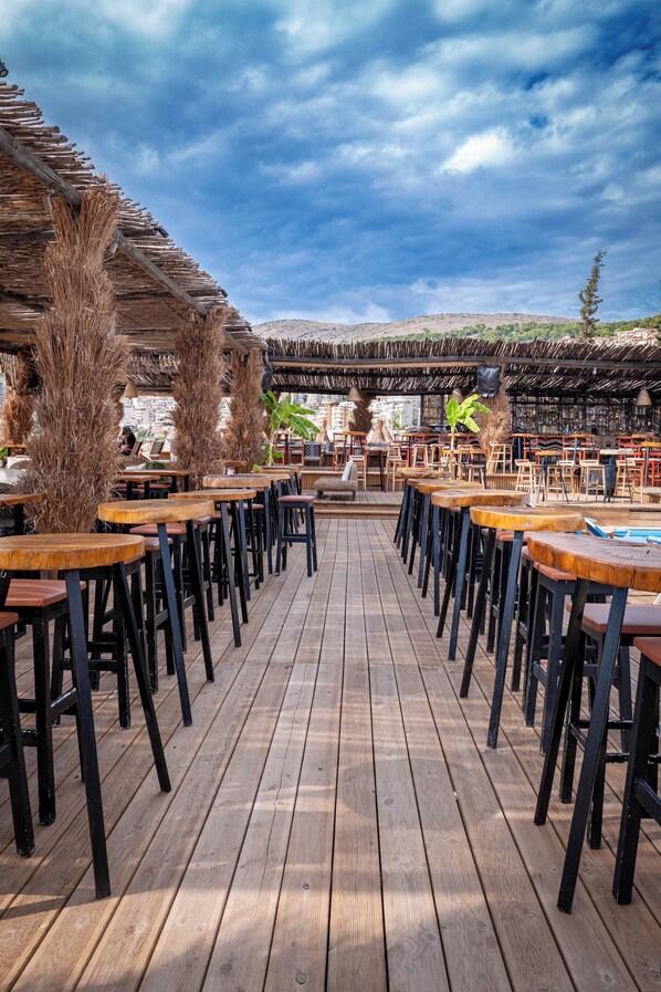 An outdoor dining area with wooden tables and chairs, a thatched roof, and a view of a mountain in the background. - Grand Hotel Saranda (Sarandë)