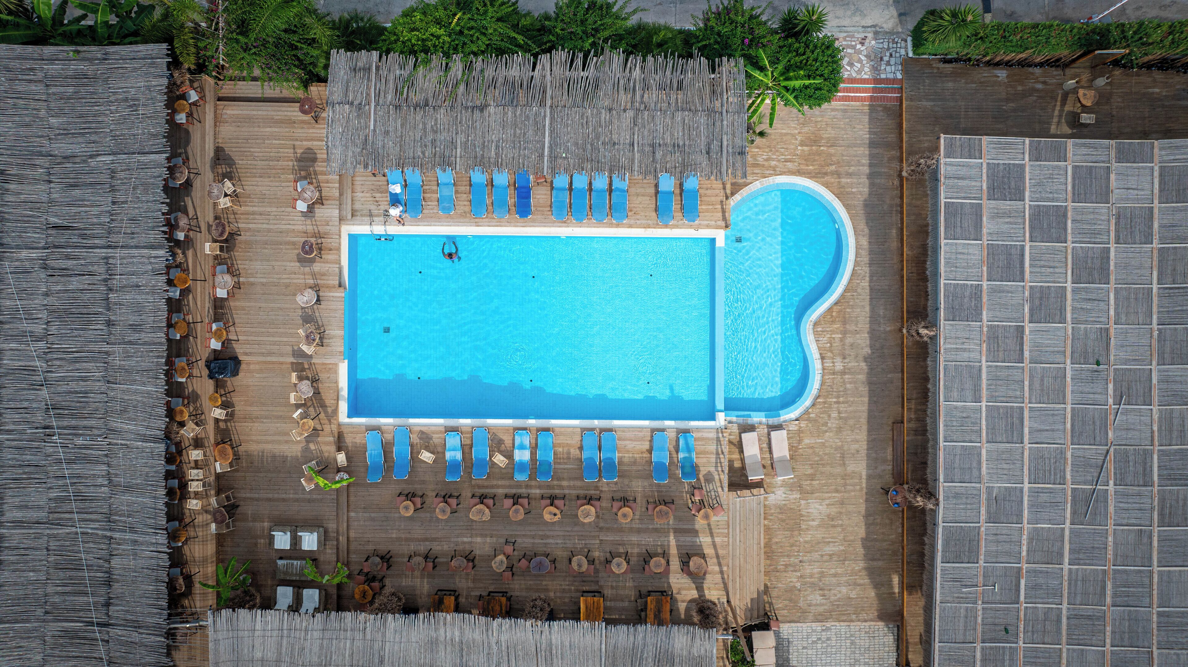 Vista aérea de uma área de piscina com várias espreguiçadeiras e mesas, cercada por um deck de madeira e uma cerca de bambu.