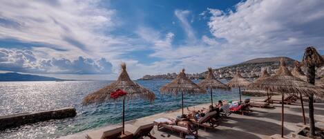 A beachside area with lounge chairs and thatched umbrellas, overlooking a body of water and a distant cityscape.