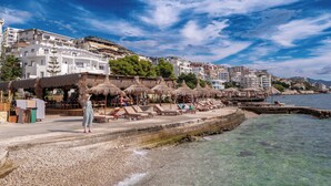 A coastal hotel with beachside lounge chairs and umbrellas, a pebble beach, and buildings in the background. - Grand Hotel Saranda (Sarandë)