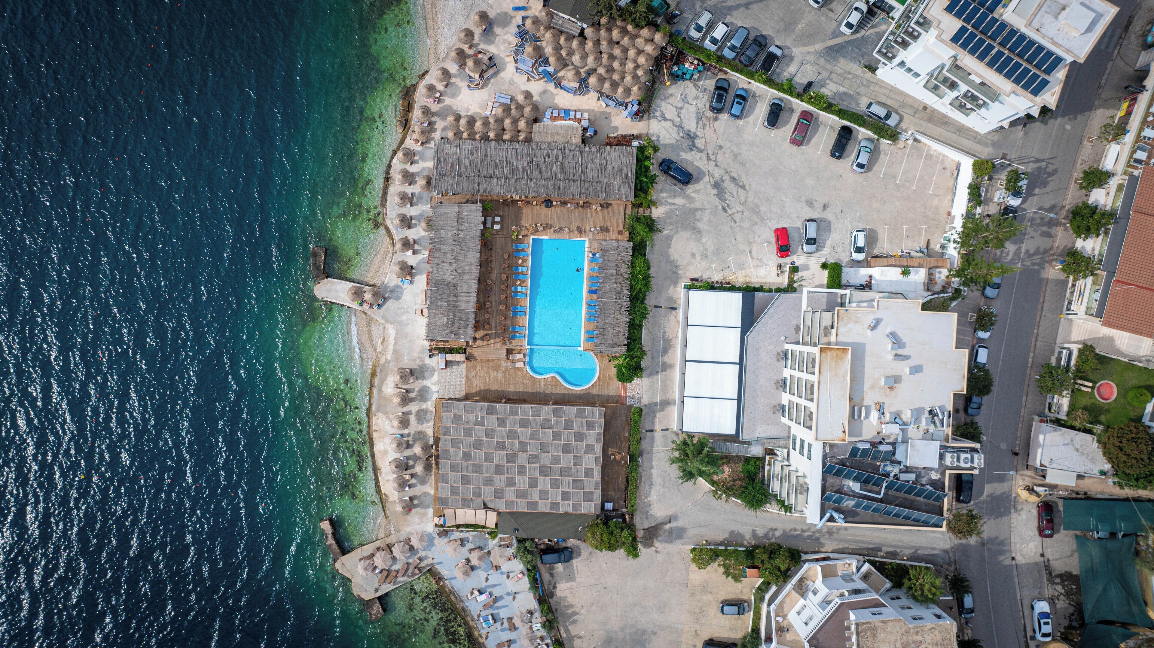 Vista aèria d'un hotel a la costa, amb piscina, seients al costat de la platja i una zona d'aparcament.
