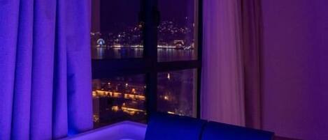 Una habitación con cama, ventana y vista a un paisaje urbano nocturno.