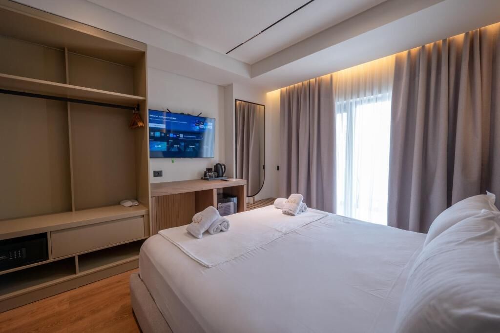 A hotel room with a large bed, a TV mounted on the wall, a desk with a computer, and a wardrobe.