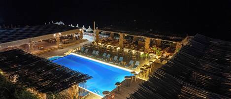 A nighttime view of a resort with a swimming pool, lounge chairs, and thatched-roof structures.