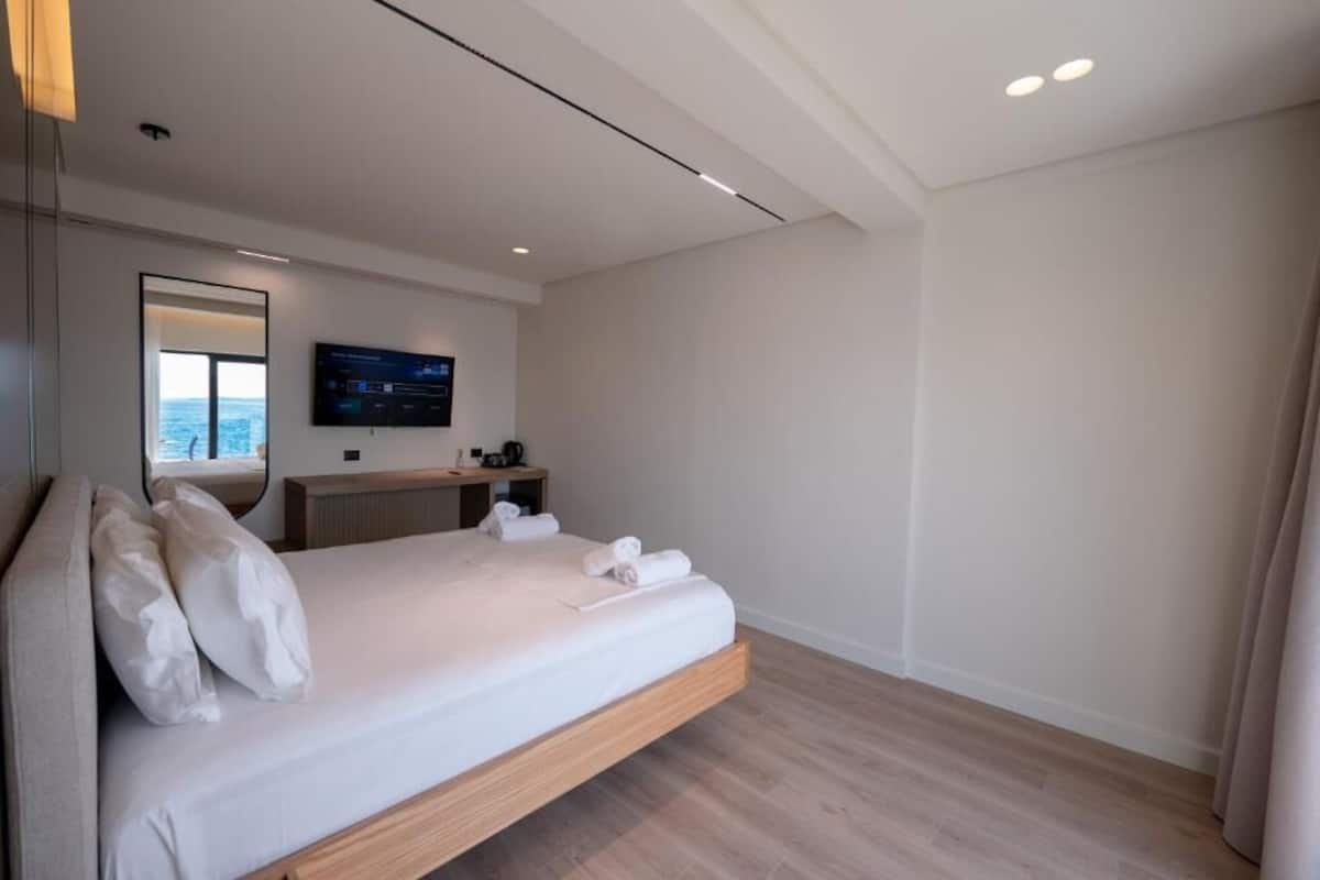 A modern hotel room with a bed, a TV, a bedside table, and a view of the sea.