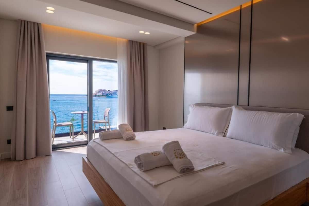 A modern hotel room with a large bed, a balcony with a view of the sea, and a table with chairs outside.