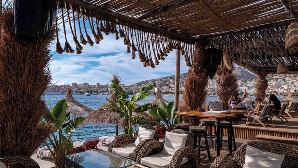 A seaside restaurant with wicker furniture, a glass table, and a view of the city and sea. - Grand Hotel Saranda (Sarandë)