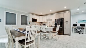 A modern kitchen with white cabinetry, a black refrigerator, and a dining area with white chairs and a wooden table. - 2 Acre 4BR Coastal Hideout: Heated Salt Pool Games (Bradenton)