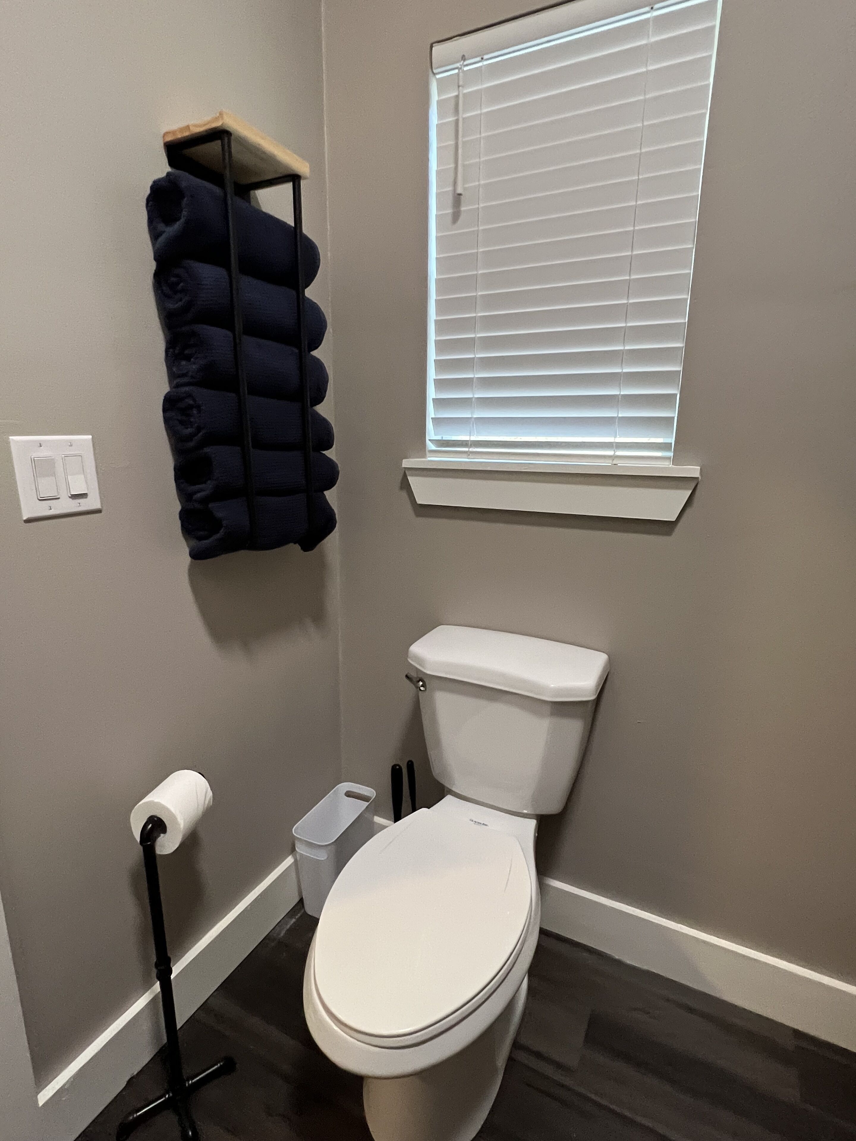Een badkamer met toilet, een handdoekrek met opgevouwen handdoeken, een raam met luxaflex en een toiletrolhouder.