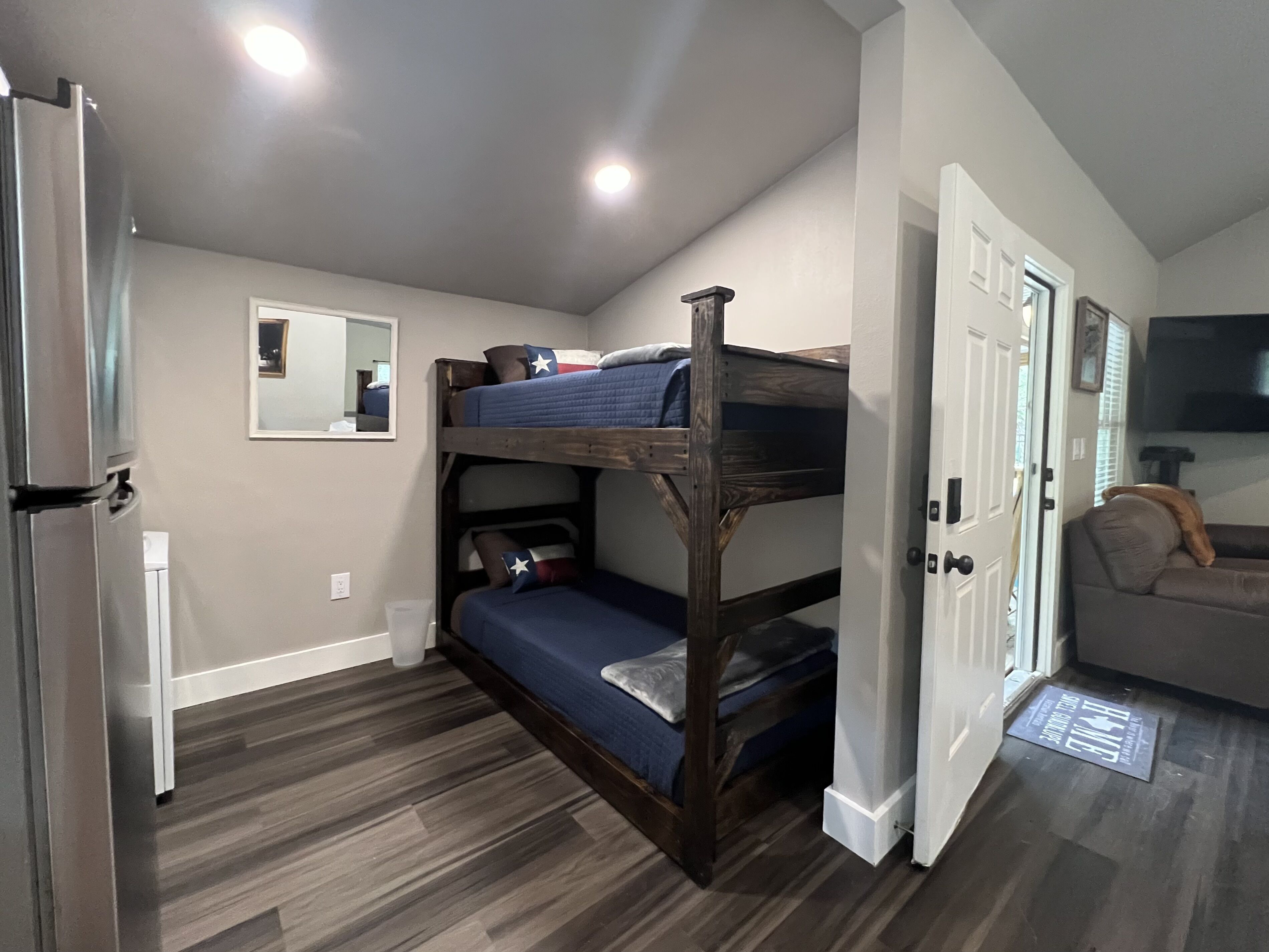 A compact room with a bunk bed, a refrigerator, a door leading to another room, and a mirror on the wall.