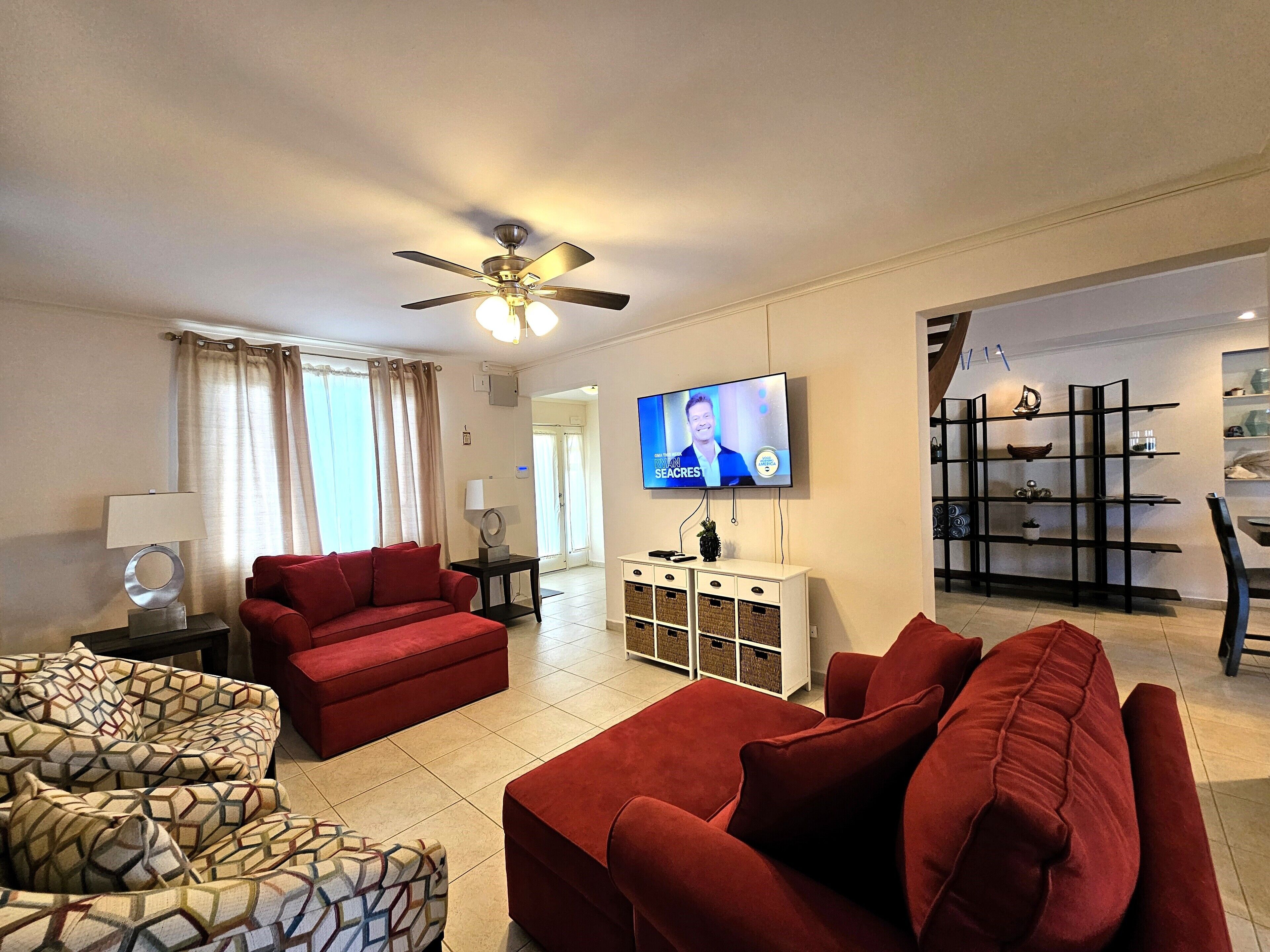Ein Wohnzimmer mit einem roten Sofa, einem gemusterten Sessel, einem an der Wand montierten Fernseher und einem Bücherregal.