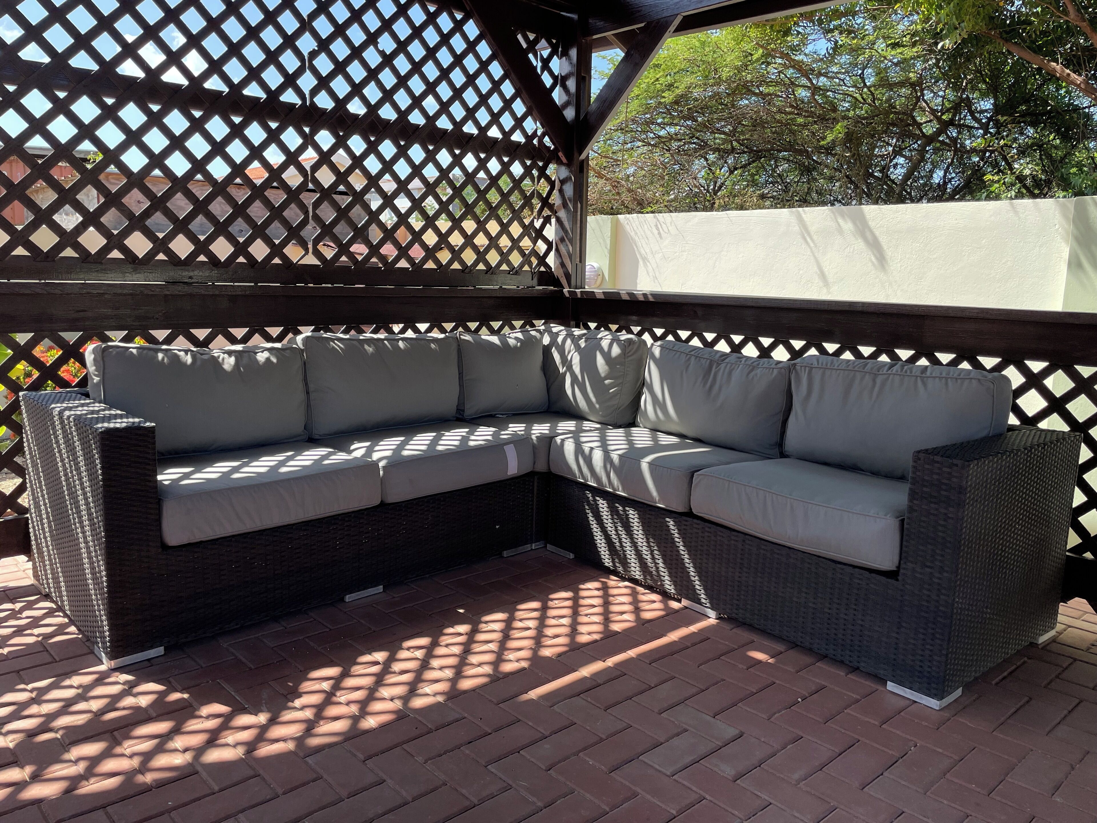 A corner sectional sofa with cushions, placed on a brick patio under a wooden pergola.