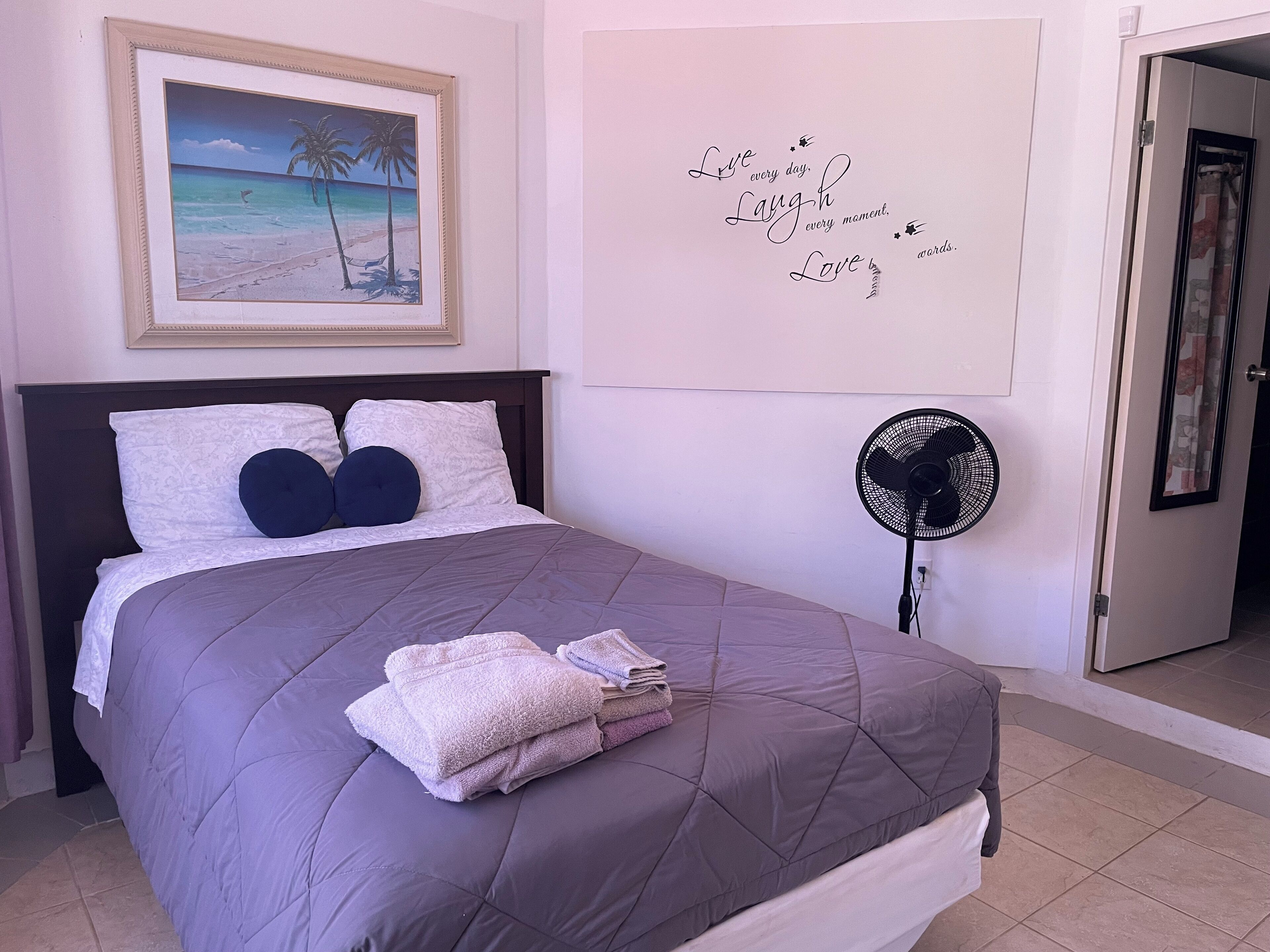 A neatly made bed with a grey quilt and folded towels, a framed picture of a beach scene, and a wall with inspirational words.