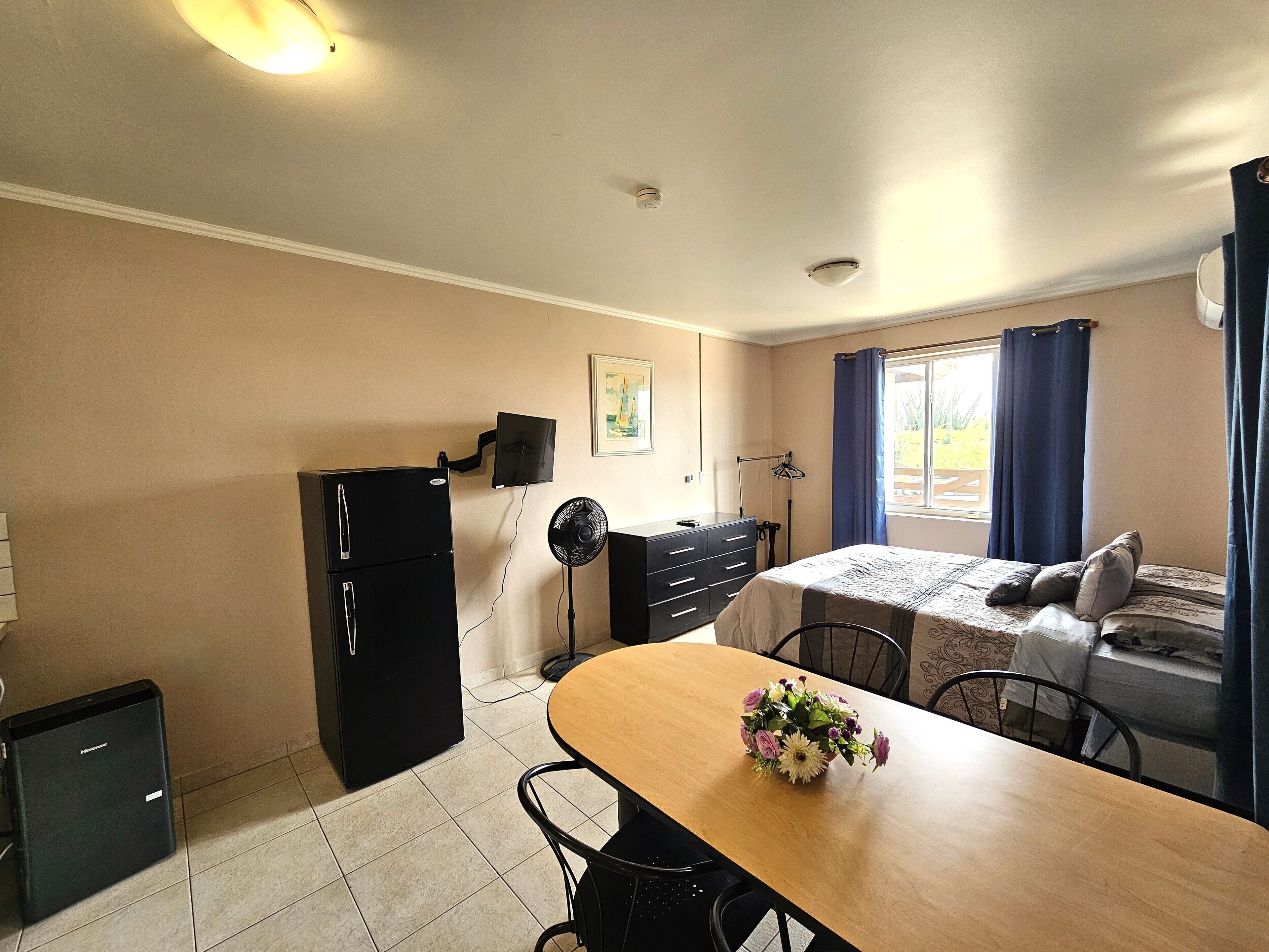 A compact room with a bed, a small table with a flower vase, a refrigerator, a dresser, a desk with a computer monitor, and a window with curtains.