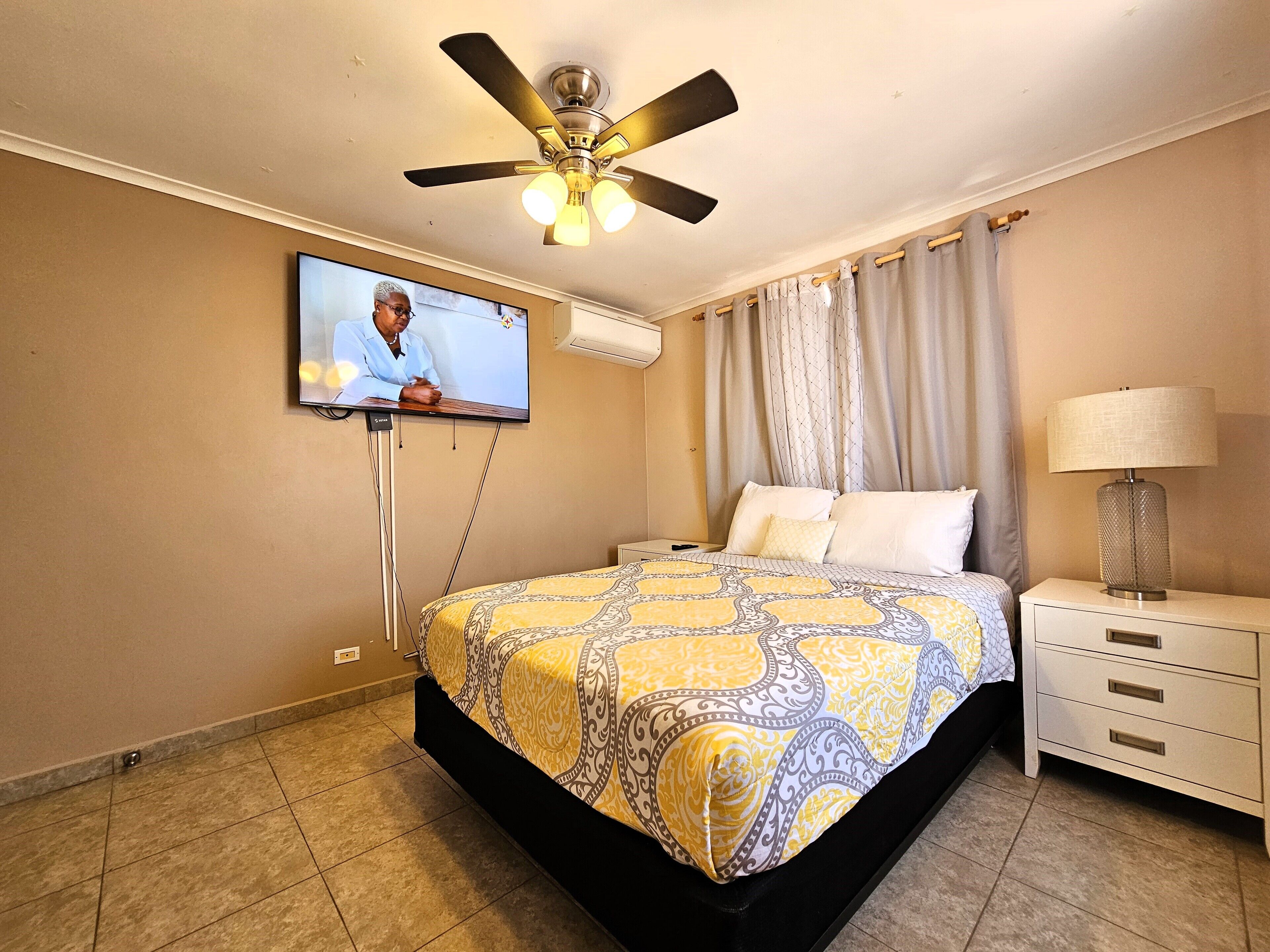 Une chambre d’hôtel avec un lit, un téléviseur fixé au mur, un ventilateur de plafond, une table de chevet avec une lampe et une fenêtre avec des rideaux.