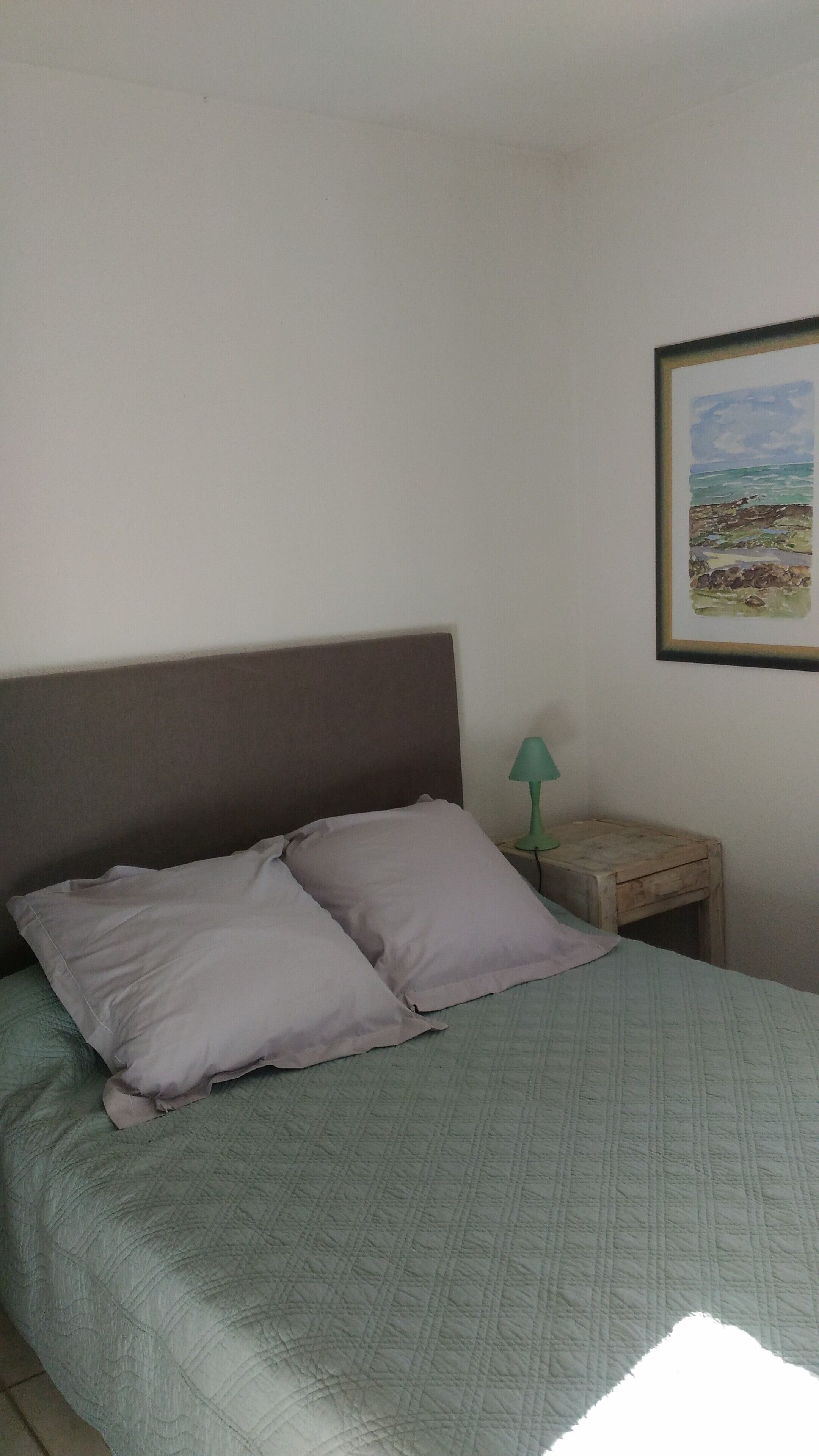Una cama con colcha verde, dos almohadas, una mesita de noche de madera con una lámpara verde y un cuadro en la pared.