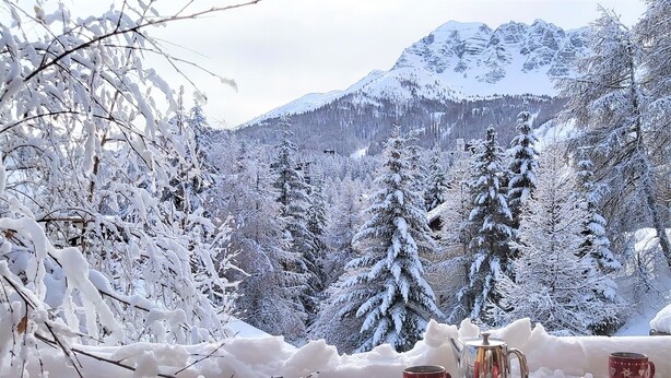 A wooden table with red cups and a teapot on a balcony overlooking a snowy mountain landscape.