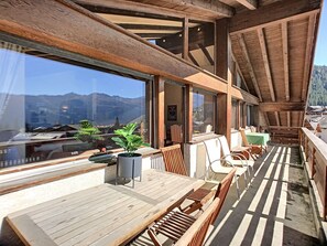 Um deck de madeira com mesa e cadeiras, grandes janelas proporcionando vista para uma paisagem montanhosa.