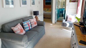 Ett modernt vardagsrum med en grå soffa, två inramade affischer med marint tema, ett litet bord med en lampa och en tv placerad på ett träställ.